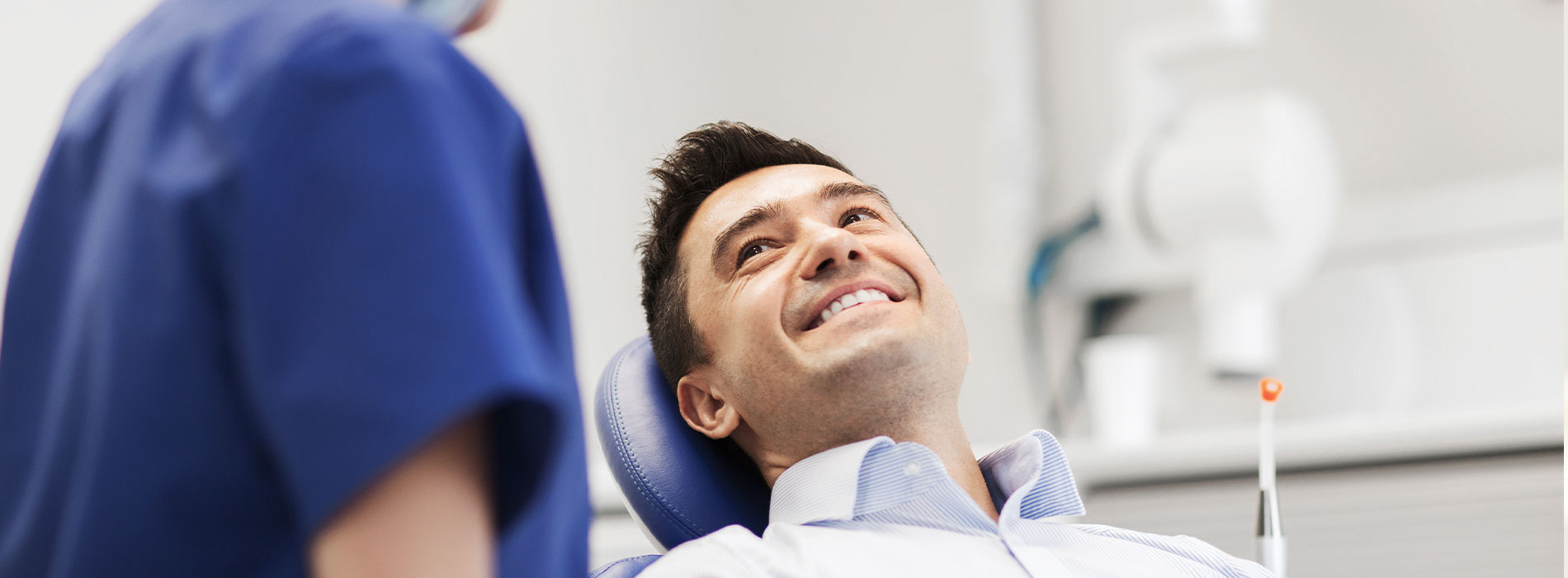 The image depicts a dental patient seated in a chair with a smiling expression, receiving care from a dental professional who appears to be examining their teeth.