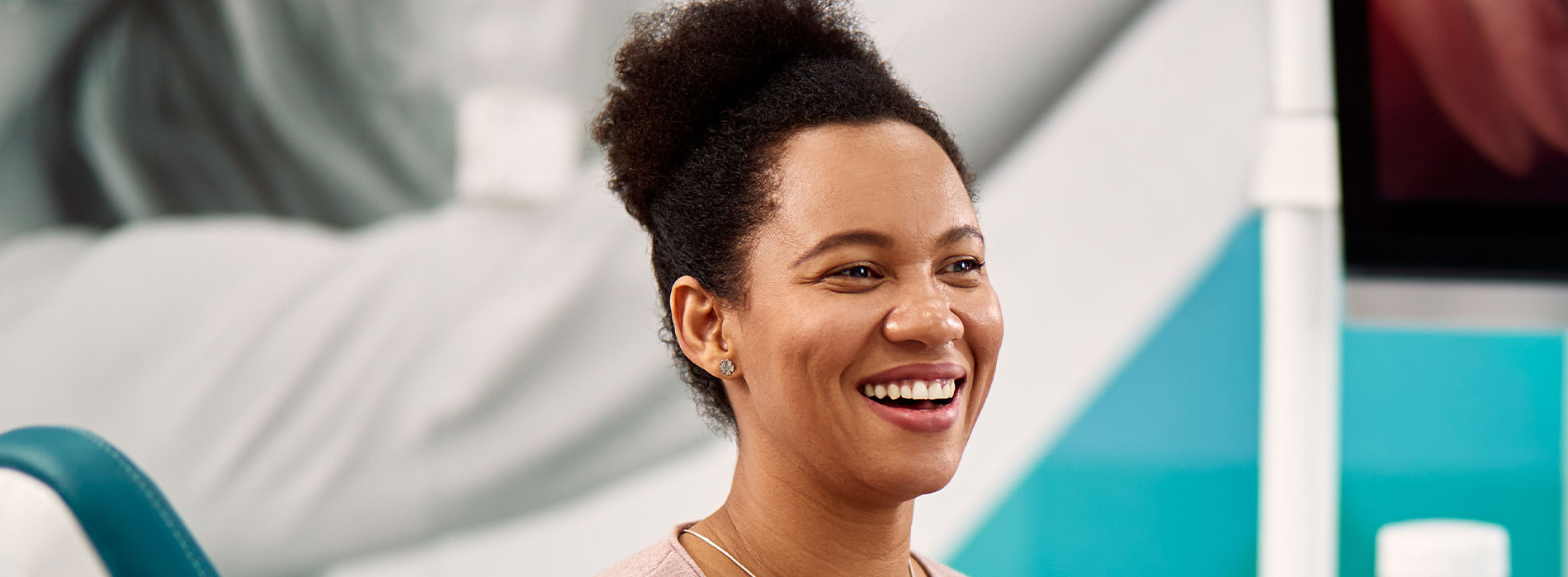 The image shows a woman smiling at the camera with her hands behind her back, wearing a light-colored top, against a blurred background that suggests an indoor setting.