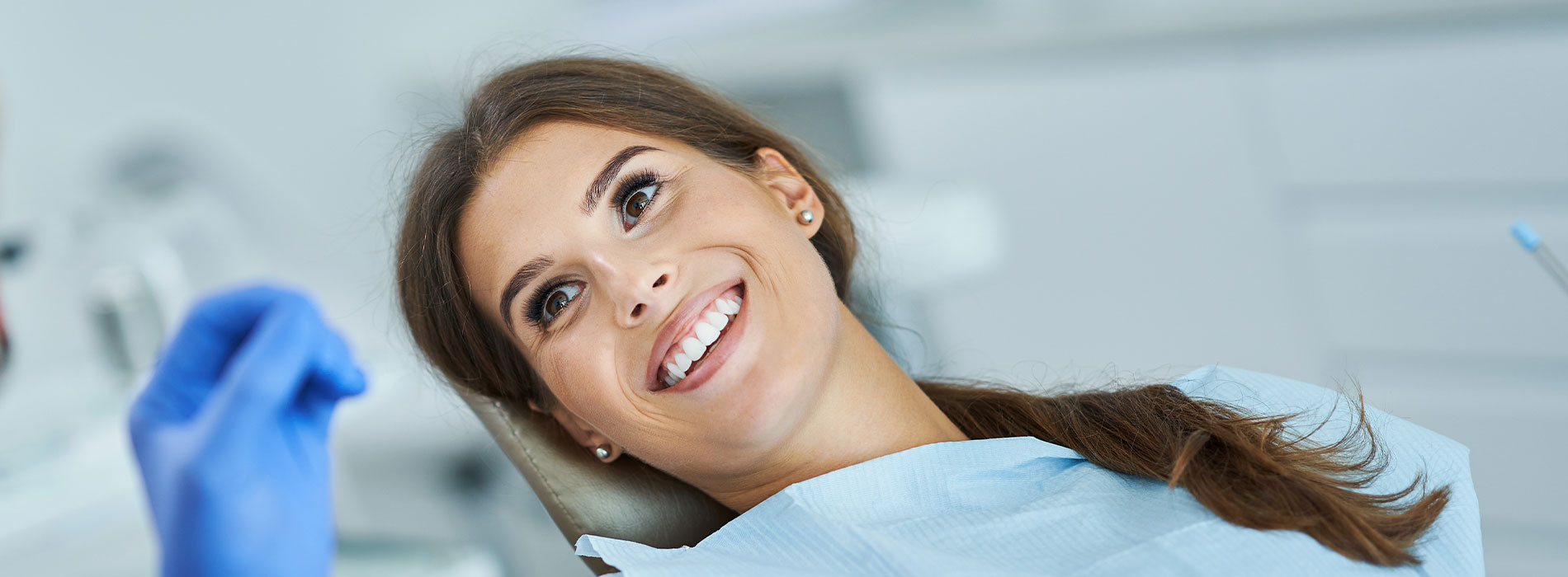 A smiling woman wearing a blue surgical mask and seated at a dental chair with medical equipment behind her.