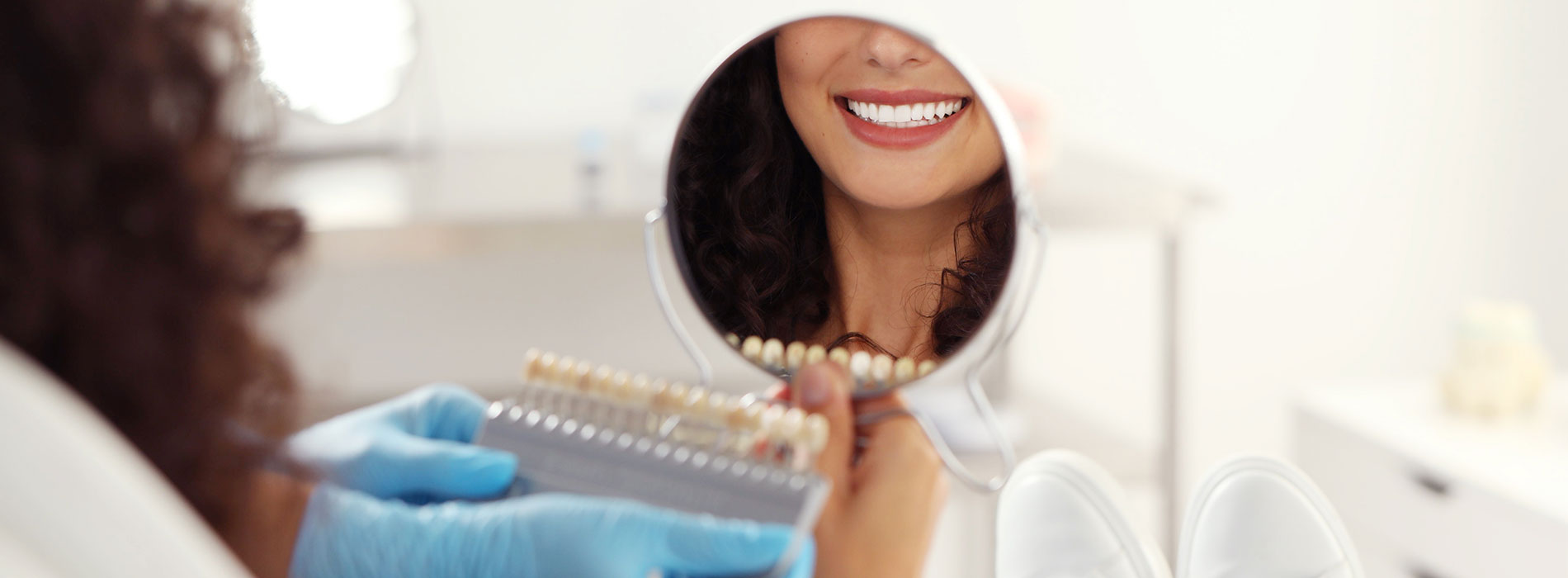 A woman with a bright smile is seen holding a tray of dental implants, standing in front of a mirror in what appears to be a dental office setting.