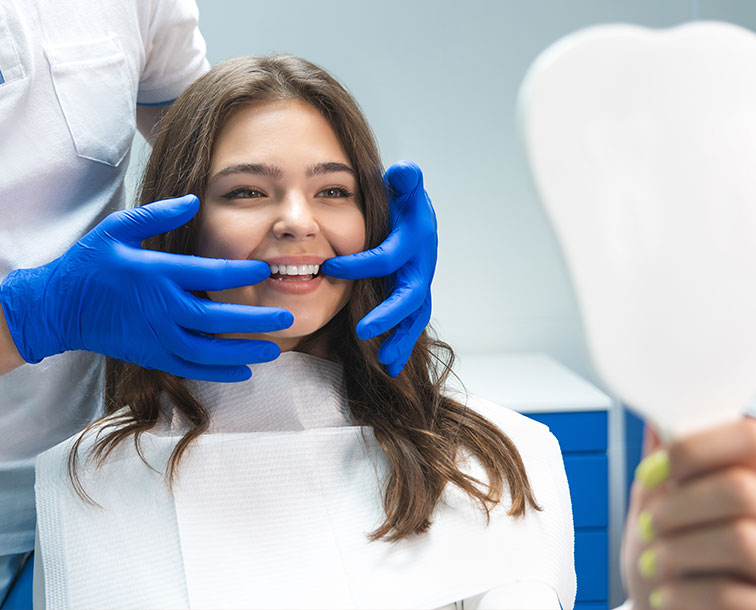 The image shows a woman seated in a dental chair, smiling at the camera, with her mouth open as if receiving dental care. A dental professional is holding up a mirror to show the woman s teeth, wearing blue gloves and a face mask.
