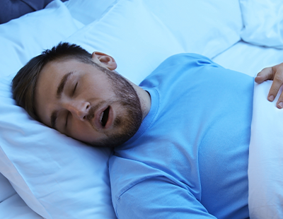 The image shows a man lying in bed with his eyes closed, appearing to be asleep, with a pillow under his head and a white sheet covering him.