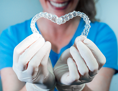 A woman wearing gloves holds up a transparent plastic heart-shaped object, possibly an orthodontic appliance, against a blue background.