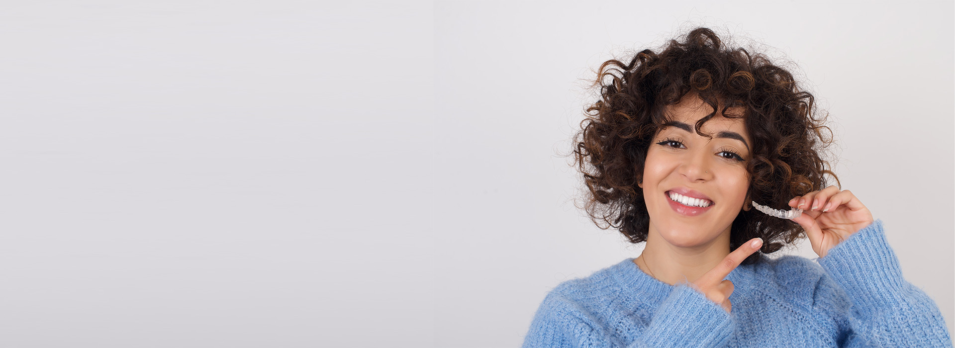 A smiling woman with curly hair stands against a white background, holding a smartphone.