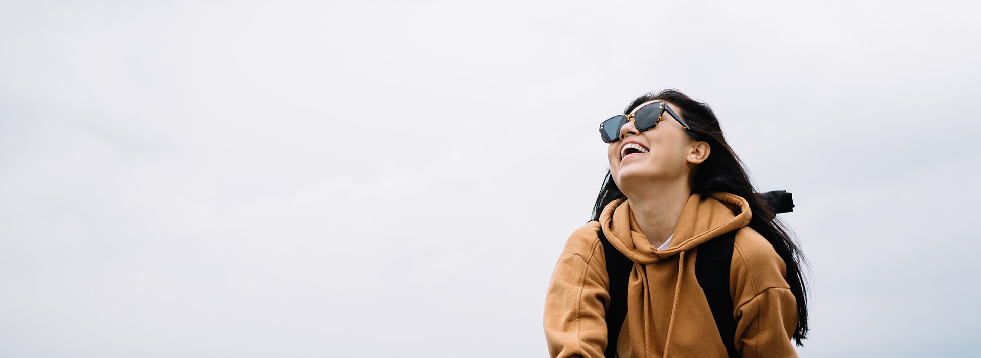 A person standing outdoors with sunglasses on, smiling, and wearing a backpack, set against a cloudy sky.