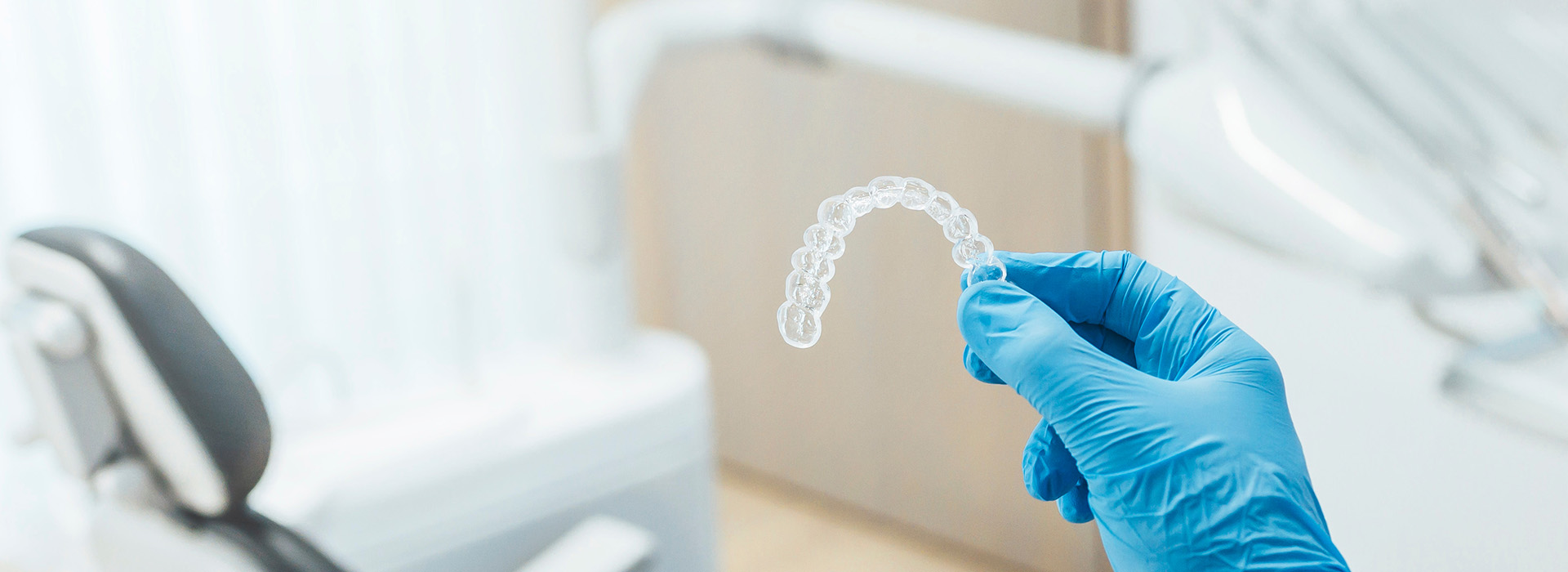 The image shows a close-up view of a transparent dental impression being held by a person wearing blue gloves, with a blurred background featuring dental equipment and a white coat.