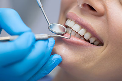 In the image, there s a person receiving dental treatment with a smile on their face, wearing a blue surgical mask and having a dental tool applied by a dental professional in a clinical setting.