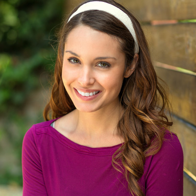 A smiling woman with long hair, wearing a purple top and headband, stands against a wooden fence.