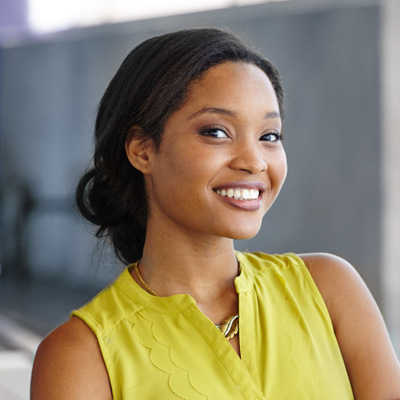 A woman with dark hair smiles at the camera, wearing a yellow top and standing against a backdrop that includes a wall and a railing.