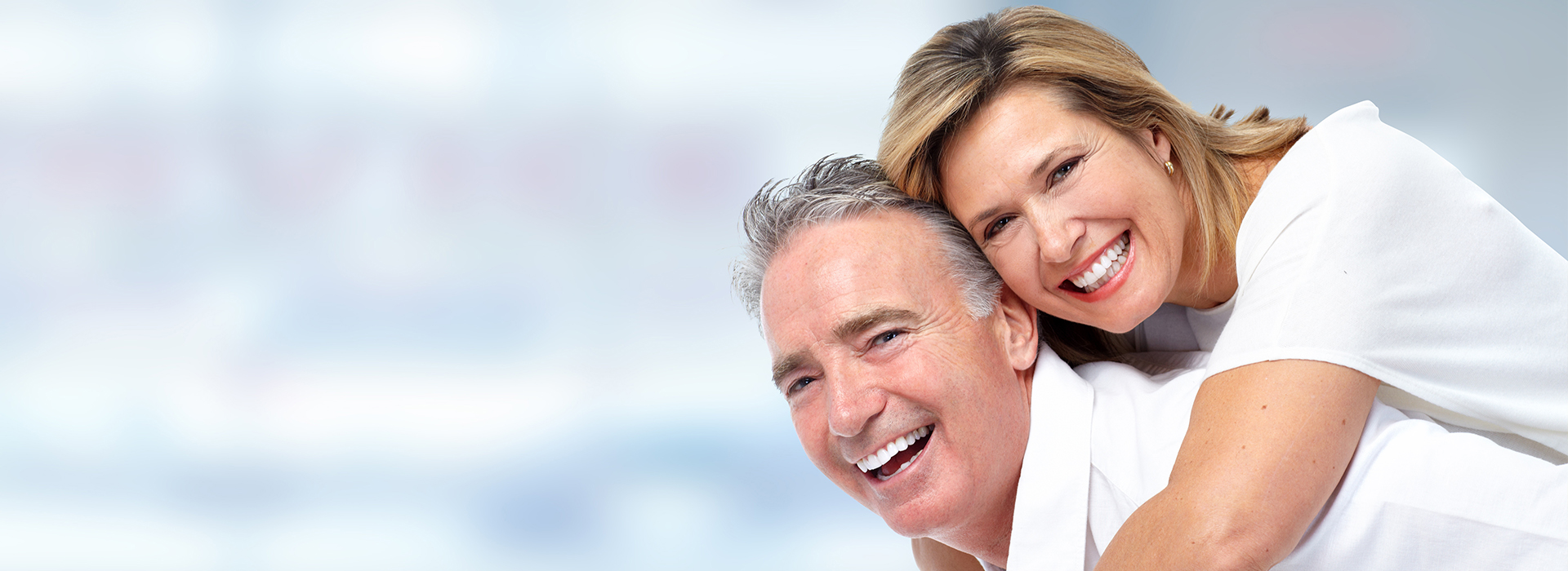 The image shows two people, an older man and woman, embracing each other with smiles on their faces, set against a blurred background featuring a blue sky.