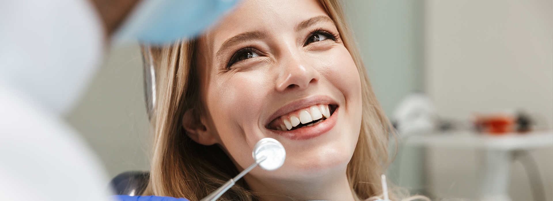 This is a photograph featuring a woman sitting in front of a dental chair with a dental professional behind her, smiling at the camera while looking content.