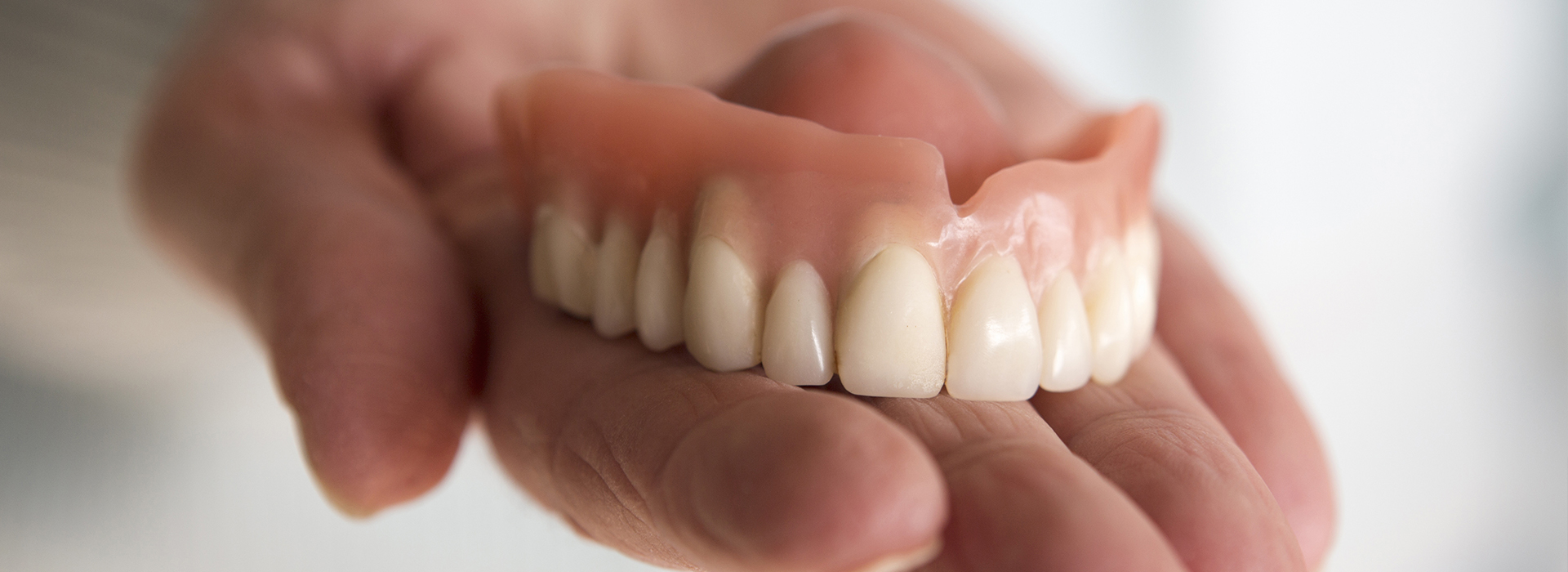 The image shows a person holding a set of dentures with their left hand against a light background.