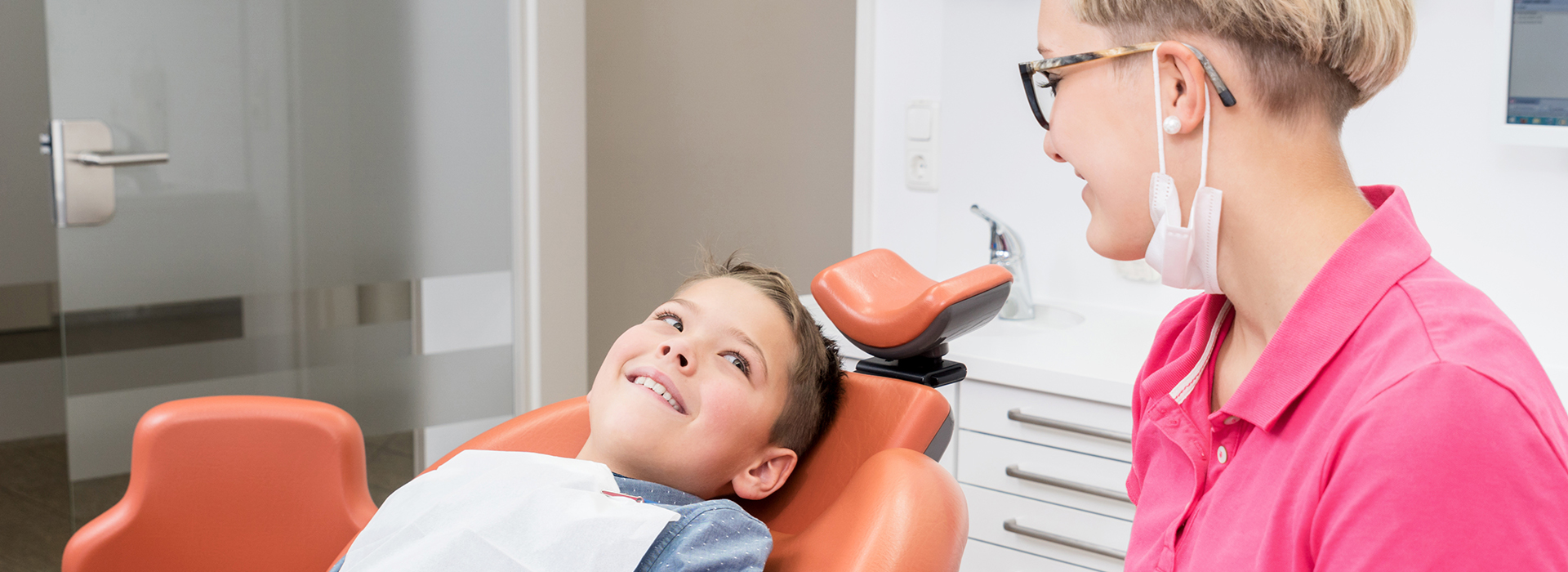 A child sitting in a dental chair, with a dental professional standing beside him, smiling and engaging with the child.