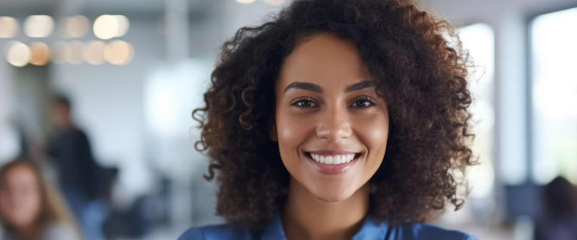 The image shows a smiling woman with curly hair, wearing a professional dark top, standing in an office environment with blurred background figures.