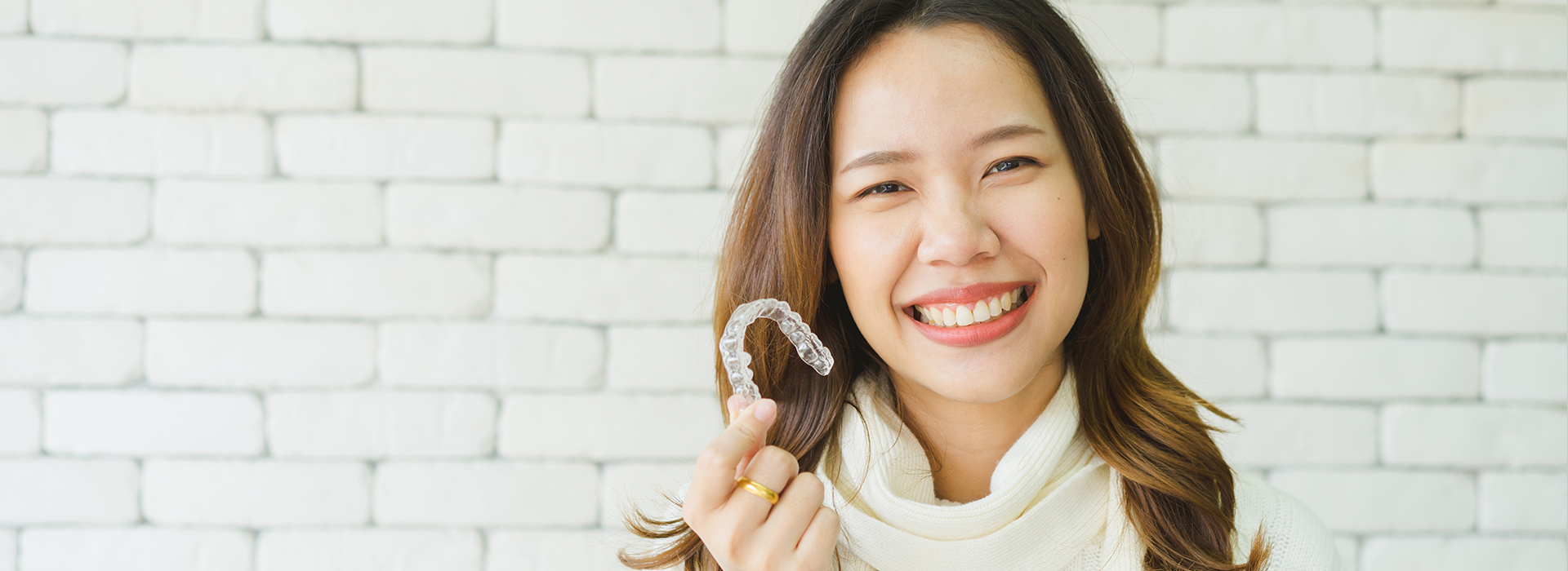 A smiling woman wearing a ring, set against a brick wall background.