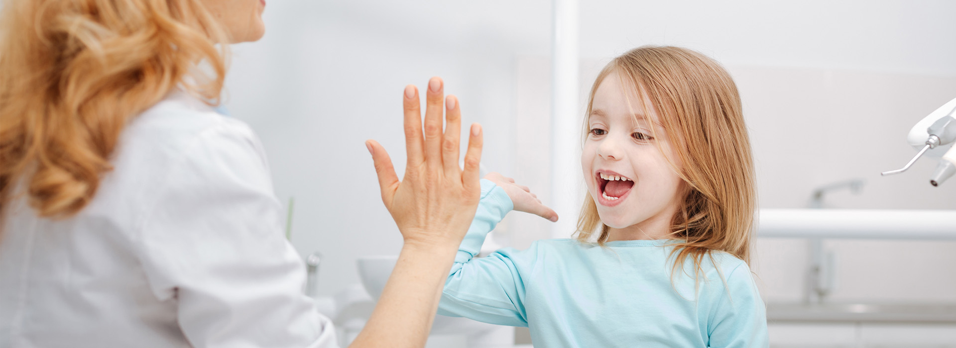 The image shows a woman and a young girl with their hands raised towards each other in a room with a sink and mirror, likely during a dental visit.