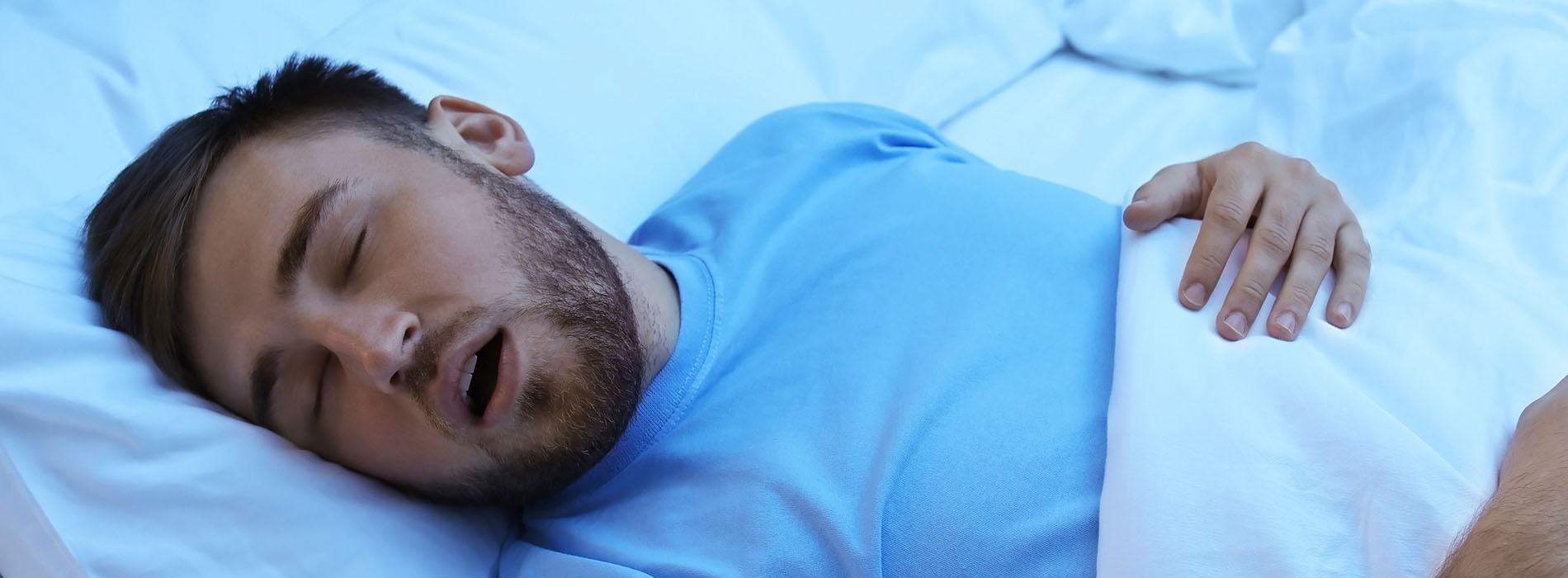 A bearded man in a blue shirt sleeping on his back with eyes closed in bed.