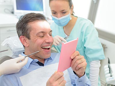 A man sitting in a dental chair with a smile on his face, holding up a pink card, surrounded by dental professionals who are examining him.