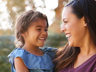 A young girl smiling at her mother, with both sharing a joyful moment outdoors during daylight.