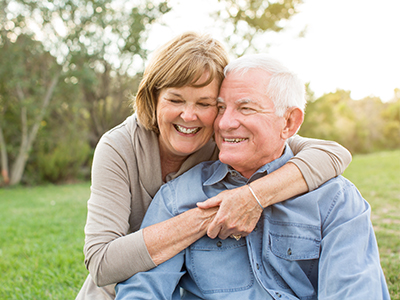 The image shows an elderly couple, a man and a woman, sharing a warm embrace while sitting on grass outdoors during daylight.