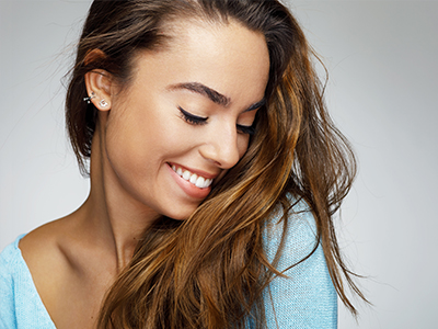 The image shows a young woman with long brown hair smiling gently at the camera while looking downwards. She has fair skin, wears a light blue top, and her expression conveys a sense of warmth and contentment.