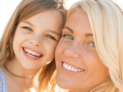 The image shows a woman and a young girl smiling at the camera  they appear to be enjoying a sunny day outdoors, with the woman likely being the child s mother.