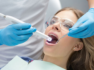 The image shows a person receiving dental care with a dental hygienist using an electric toothbrush to clean their teeth while they are seated in a dental chair, wearing protective gloves and blue surgical attire.