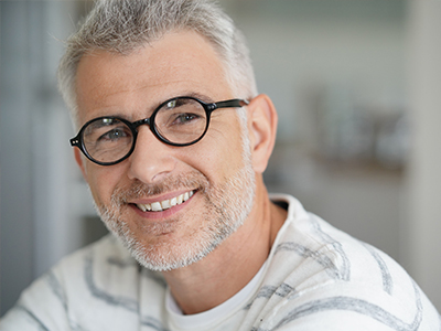 The image features a man with gray hair, wearing glasses and a white shirt with a patterned collar, smiling at the camera.