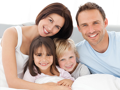 The image shows a family of four people lying on a bed with smiles on their faces, likely enjoying a moment together.