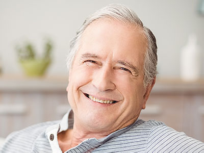 The image features an elderly man with gray hair, smiling and looking directly at the camera. He has white hair and appears to be in a relaxed posture, possibly seated in a home environment.