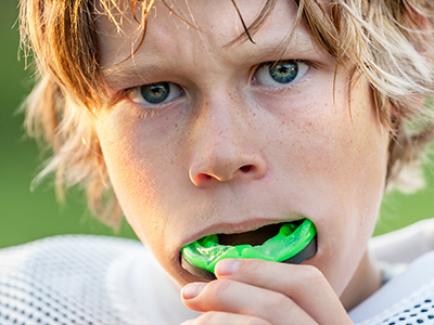 A young boy with blonde hair, wearing a white jersey, holding a green toothbrush in his mouth while making an unusual face.