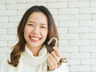 A woman with a bright smile holding up a toothbrush, set against a brick wall background.